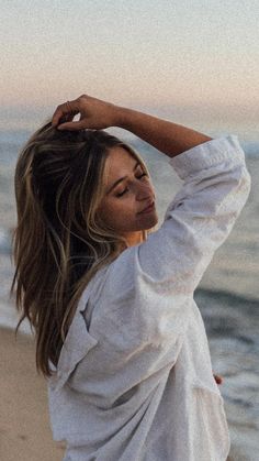 a woman standing on top of a beach next to the ocean holding her hair up