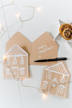 three gingerbread house christmas cards on a table with a pen and orange slice next to them