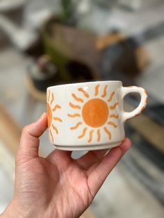 a hand holding an orange and white coffee cup with the sun painted on it's side