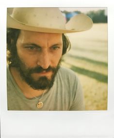 a man with a beard wearing a hat and looking at the camera while standing outside