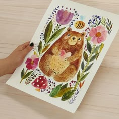 a person holding up a card with a bear and bee on it, surrounded by flowers