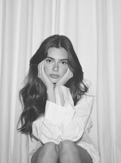 a black and white photo of a woman with her hands on her face sitting in front of a curtain