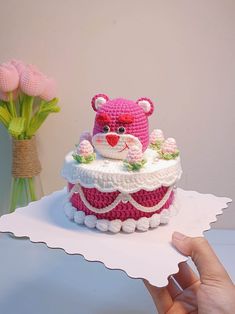 a crocheted teddy bear on top of a cake with flowers in the background