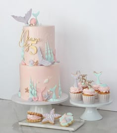 there is a pink and blue cake with mermaid decorations on the top, along with cupcakes