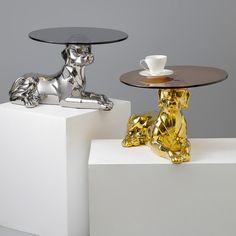 two glass tables sitting next to each other on top of white pedestals and one has a gold dog figurine