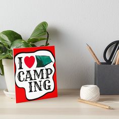 i love camp ing art board print on wood with scissors and pencils next to potted plant