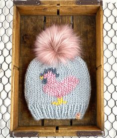 a knitted hat with a pink pom - pom sitting in a wooden box