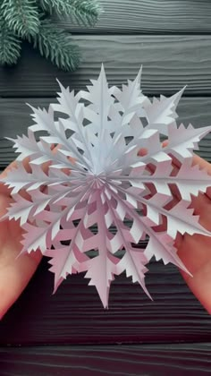 someone is holding a paper snowflake in their hand on a wooden table with pine branches