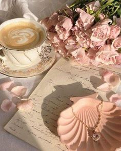 a cup of coffee next to a seashell and pink flowers on a white table cloth