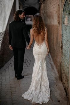 a bride and groom walking down an alleyway holding hands in their wedding gowns