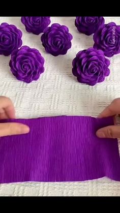 someone is making purple flowers out of yarn on a white tablecloth with pink roses in the background