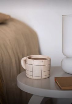 a white table with a cup on it next to a lamp and a bed in the background