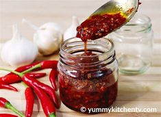 a spoon full of chili sauce being poured into a jar with red peppers and garlic on the side