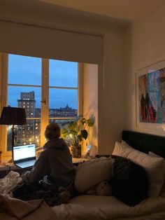 a person sitting on a bed in front of a window looking out at the city