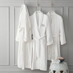 three white robes hanging on a wall next to a stool and small table with flowers