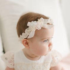 A baby in a Jessica Headband and a handmade headband with pastel floral appliqué sits on a white surface, gazing to the right. The soft lighting enhances the serene and delicate atmosphere of the scene. Christening Gowns For Boys, Pink Silk Ribbon, Christening Gifts For Boys, Flowers Pastel, Green Vines, Pearl Beading, Dress Champagne, Heirloom Gifts, Girl Christening