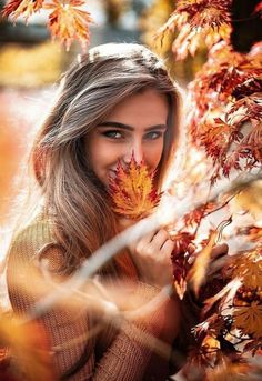 a beautiful woman holding onto some leaves in the fall season, looking at the camera