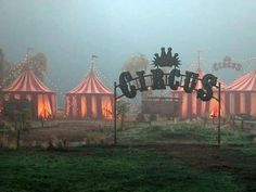 a sign that is in the grass near some tents with lights on them and trees behind it