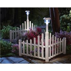 a white picket fence with a light on it in front of some flowers and trees