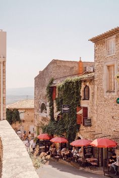 people are sitting at tables in an outdoor cafe with red umbrellas and stone buildings