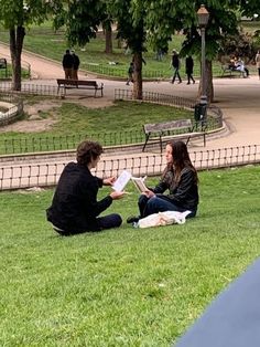 two people sitting on the grass reading books in a park with trees and benches behind them