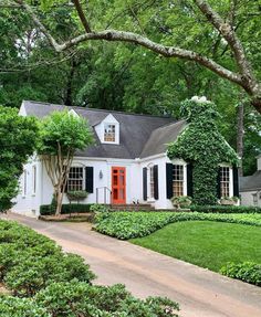 a white house with black shutters and red door surrounded by greenery in the front yard