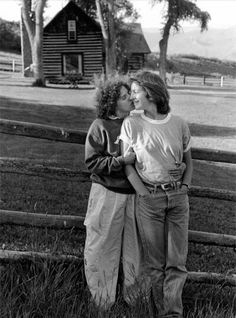 two women standing next to each other in front of a fence with a barn behind them