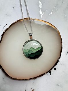 a green and white mountain pendant on a silver chain hanging from a wooden slice with a black cord