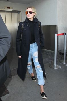 a woman in ripped jeans and black coat is walking through an airport with her hand on her hip