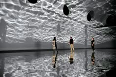 three people standing in front of a large white wall with circular lights hanging from it's ceiling