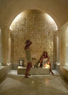 two women sitting in a stone bathtub with buckets on the side and water running down the wall