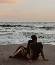 two people are sitting on the beach watching the waves come in to shore as the sun sets