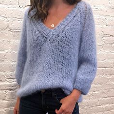 a woman standing in front of a white brick wall wearing a blue sweater and jeans