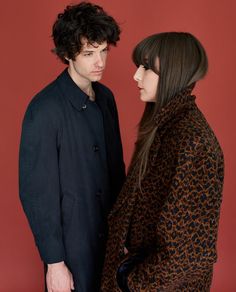 two people standing next to each other in front of a red wall and one is wearing a leopard print coat