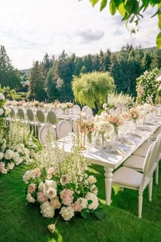 a table set up with white chairs and flowers on the grass in front of trees