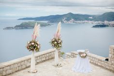 two tall vases with flowers on top of a stone wall next to water and mountains