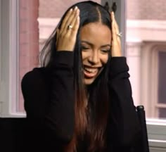 a woman with long hair smiling and holding her hands up to her head while sitting in front of a window