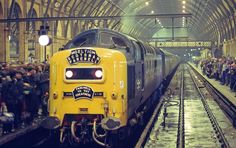 a yellow train pulling into a station with lots of people on the platform next to it