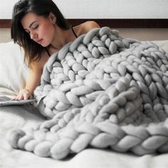 a woman laying in bed reading a book under a giant chunky knitted blanket