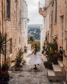 a woman in a white dress is walking down an alley way with potted plants