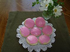 pink frosted cookies are on a doily next to a vase with white flowers