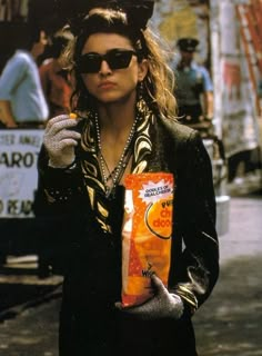 a woman wearing sunglasses and holding a bag of orange juice in her hand while walking down the street