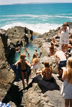 many people are sitting on the rocks near the water and one person is standing in the water