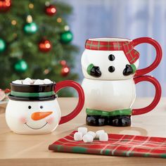 two snowman mugs sitting on top of a table next to a christmas tree