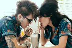 two people sitting at a table with drinks in front of their faces, one drinking from a shaker and the other eating an ice creamshake