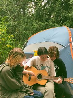 three young people sitting in front of a tent playing guitar