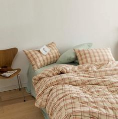 an unmade bed with plaid sheets and pillows in a white room next to a chair