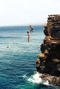 two people jumping off rocks into the ocean from a cliff face to face with each other