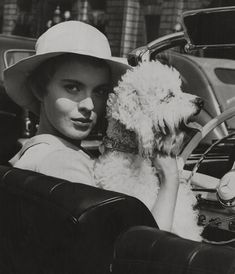 a woman sitting in a car holding a dog and wearing a hat with her hand on the steering wheel