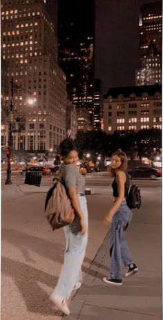 two people walking down the sidewalk in front of tall buildings at night time with lights on